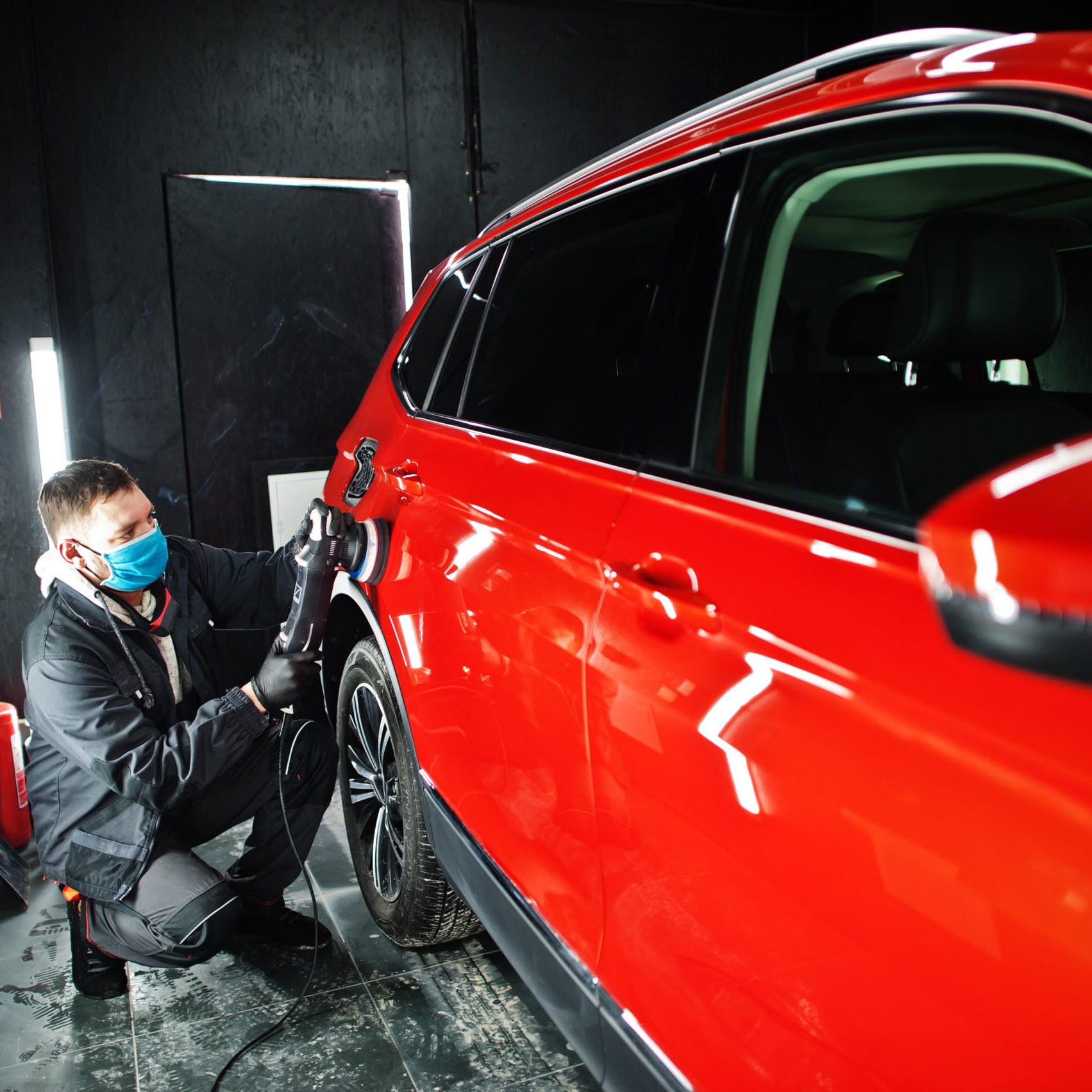 Car detailing concept. Man in face mask with orbital polisher in repair shop polishing orange suv car.